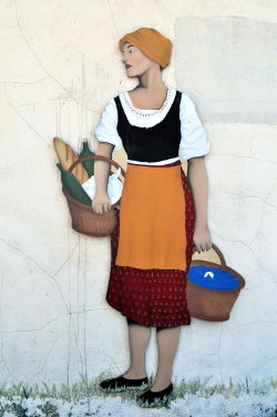 Painting of a woman in traditional dress with an orange ochre apron and headscarf and carrying two baskets of bread loaves, found painted on a wall in the Drome department.